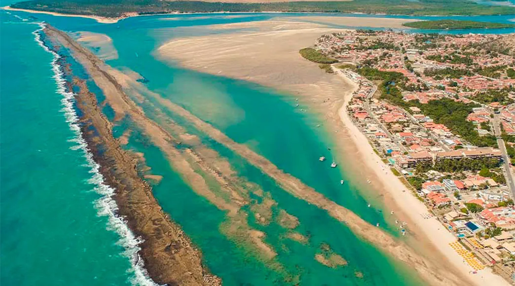 Vista aérea panorâmica de Barra de São Miguel, mostrando a longa barreira de recifes que protege a costa, a cidade à beira-mar e, ao fundo, o encontro do rio com o mar formando um grande estuário.