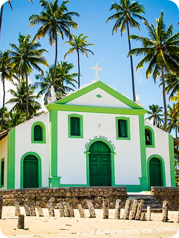 Capela de São Benedito na Praia de Carneiros, com fachada branca e verde, cercada por coqueiros, pequenos tocos de madeira na frente e areia clara sob céu azul