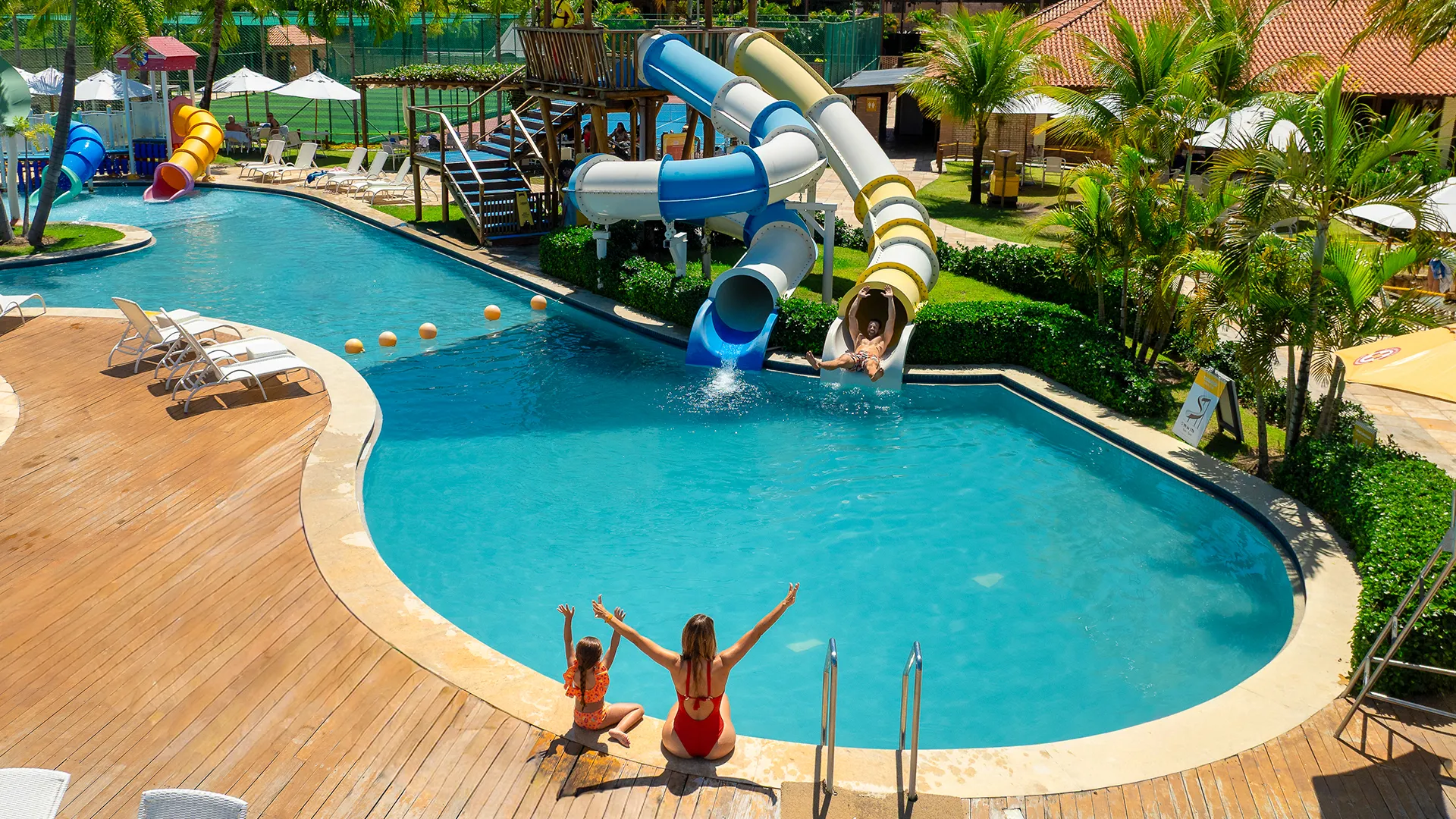 Área de lazer do Salinas Maragogi, com brinquedo de crianças com tobogã colorida na piscina. Família se divertindo, enquanto o pai desce o tobogã, mãe e filha estão sentadas na borda da piscina com os braços pra cima. Ao fundo toda a área com espreguiçadeiras, outros brinquedos, quadras, altos coqueiros verdes e estrutura do resort.