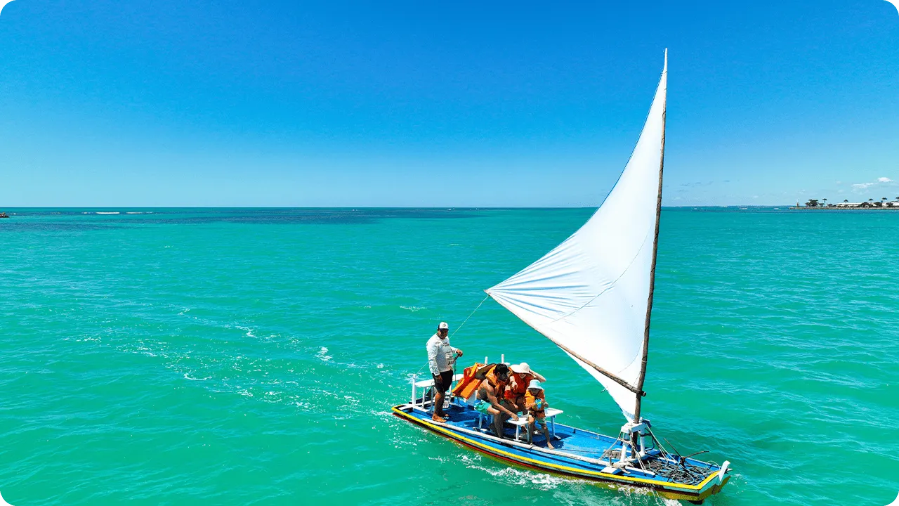 Barquinho a vela colorido navegando com o marinheiro em águas turquesa com céu azul com uma família sorrindo a bordo.