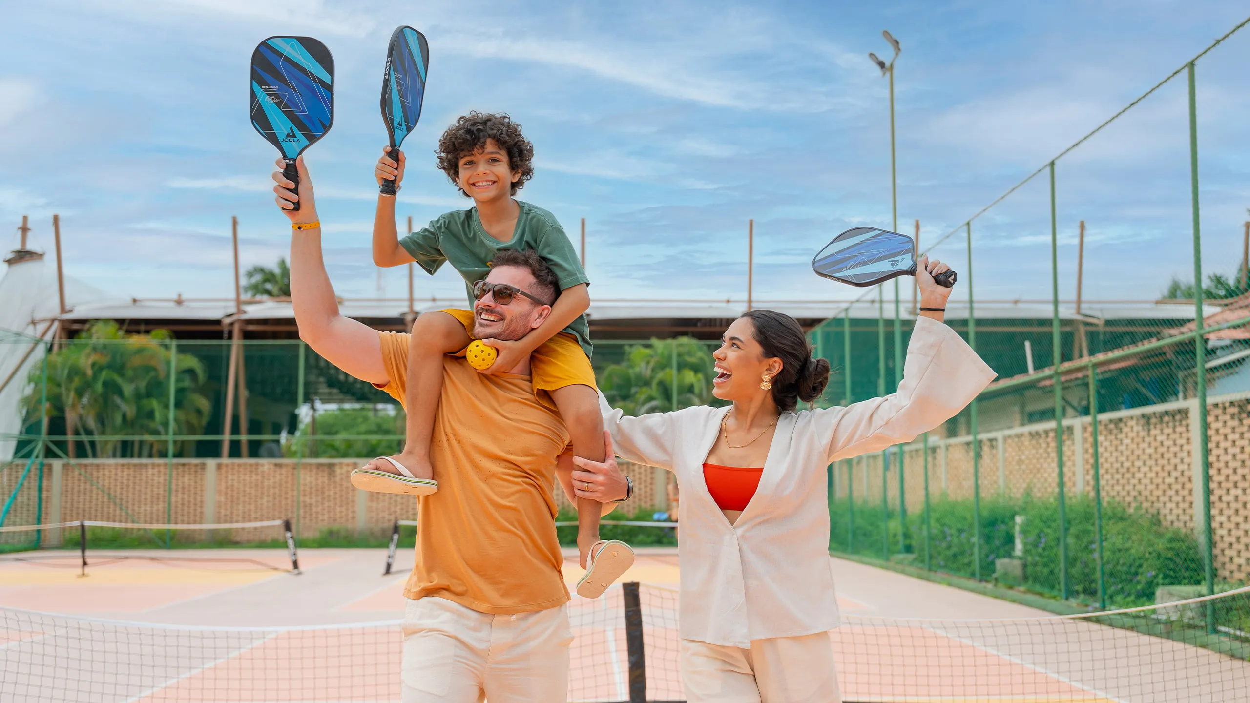Família na quadra de pickleball, com mãe, pai e filho nas costas do pai. Eles estão com raquetes e bola na mão em área de recreação do Salinas Maragogi.