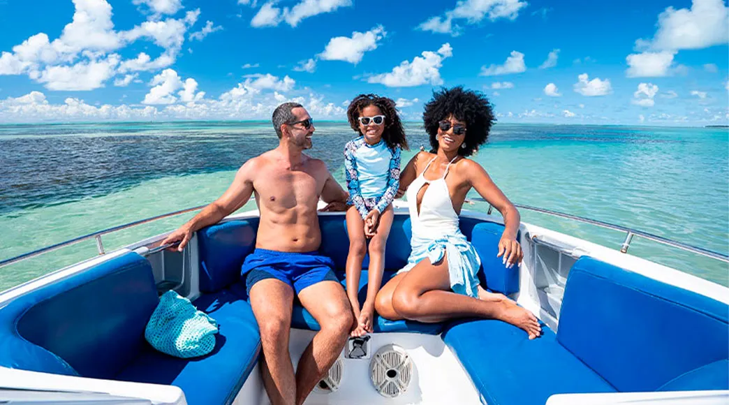 Família com pai, mãe e filha sorriem sentados na proa de uma lancha durante um passeio no mar de Paripueira. Ao fundo, o mar está calmo e o céu azul com nuvens.