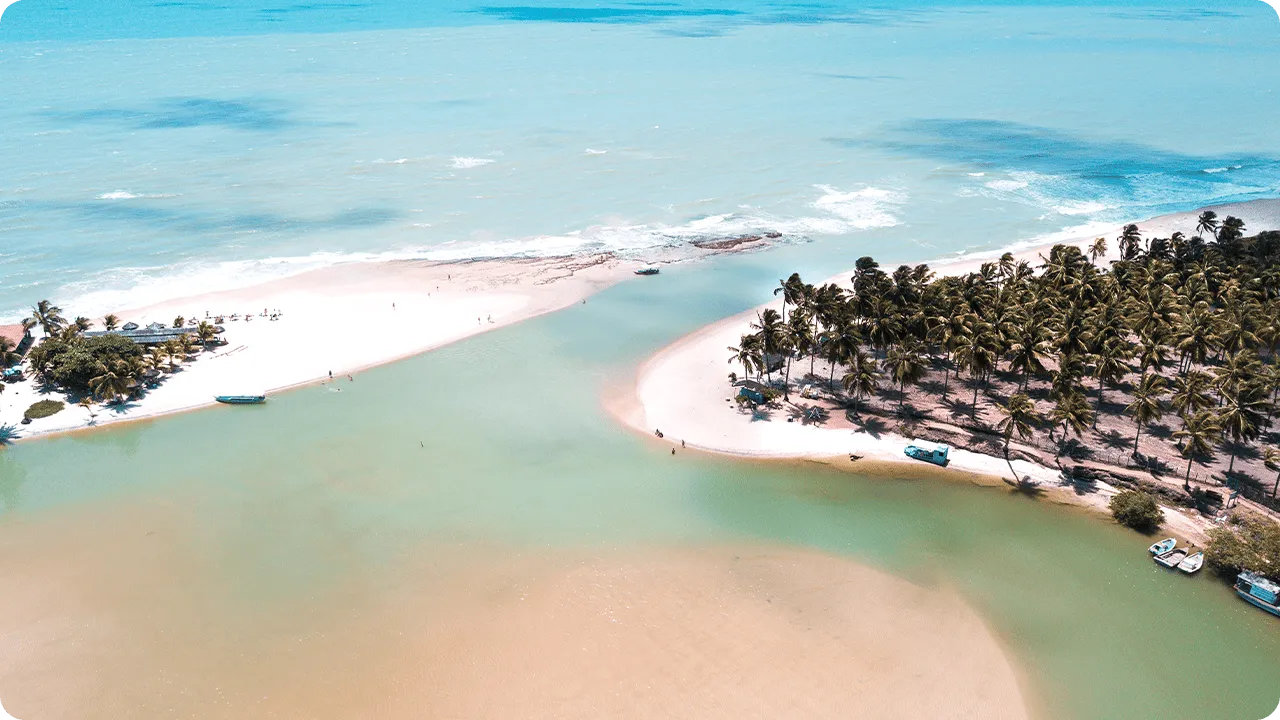 Vista aérea com encontro do rio e mar de Jequiá da Praia (Dunas de Marapé) em tons de azul e verde, faixa de areia clara em curva e um banco de areia no meio da água, coqueiros densos e barcos próximos à margem.