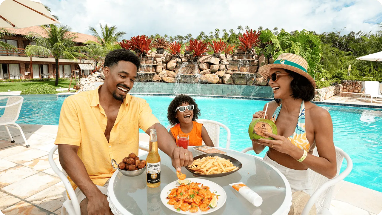 Una familia feliz con padre, madre e hija disfrutando de bocadillos y bebidas en una mesa junto a la piscina en Salinas Maragogi, con camarones, papas fritas y coco.