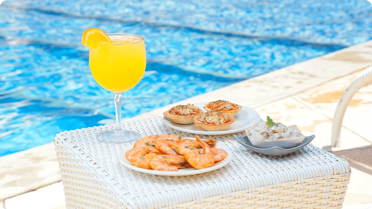 Mesa junto a la piscina con refresco de naranja y aperitivos, incluyendo camarones hervidos y mini tartas saladas, con el agua azul de la piscina de fondo.