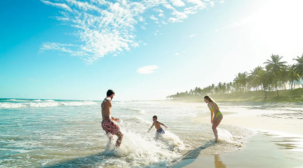 Família com pai, mãe e filho brincando nas ondas na beira do mar da Praia do Francês. O pai e o filho espirram água enquanto a mãe sorri, com coqueiros ao fundo em um dia de sol.