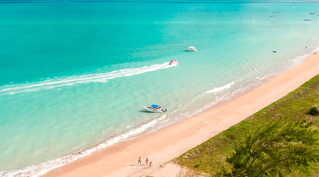Vista aérea da Praia de Burgalhau, com sua longa faixa de areia e vegetação costeira. O mar exibe tons de azul-turquesa, com uma lancha navegando em velocidade e deixando um rastro branco na água.