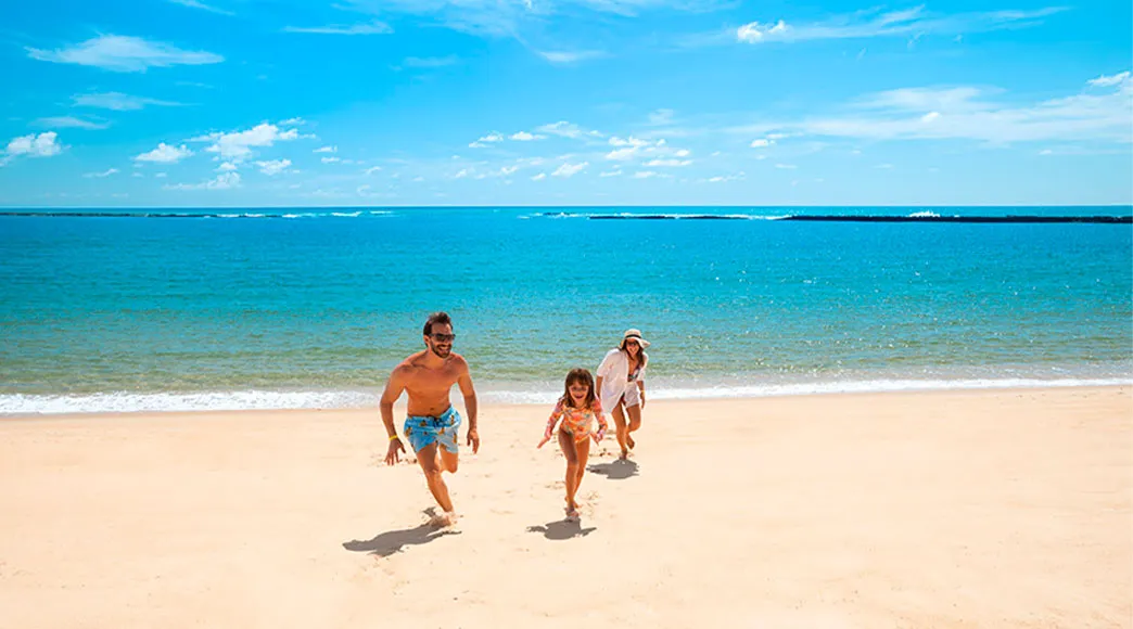 Família com pai, mãe e filha corre sorrindo na ampla faixa de areia da Praia do Gunga, com o mar calmo e azul ao fundo sob um céu ensolarado.