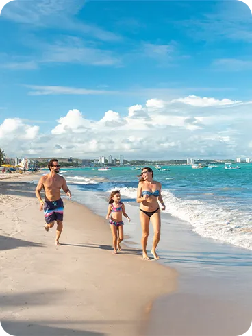 Família correndo pela areia clara da praia de Jatiúca com mar turquesa quase em seus pés, barcos ao fundo e cidade no horizonte.