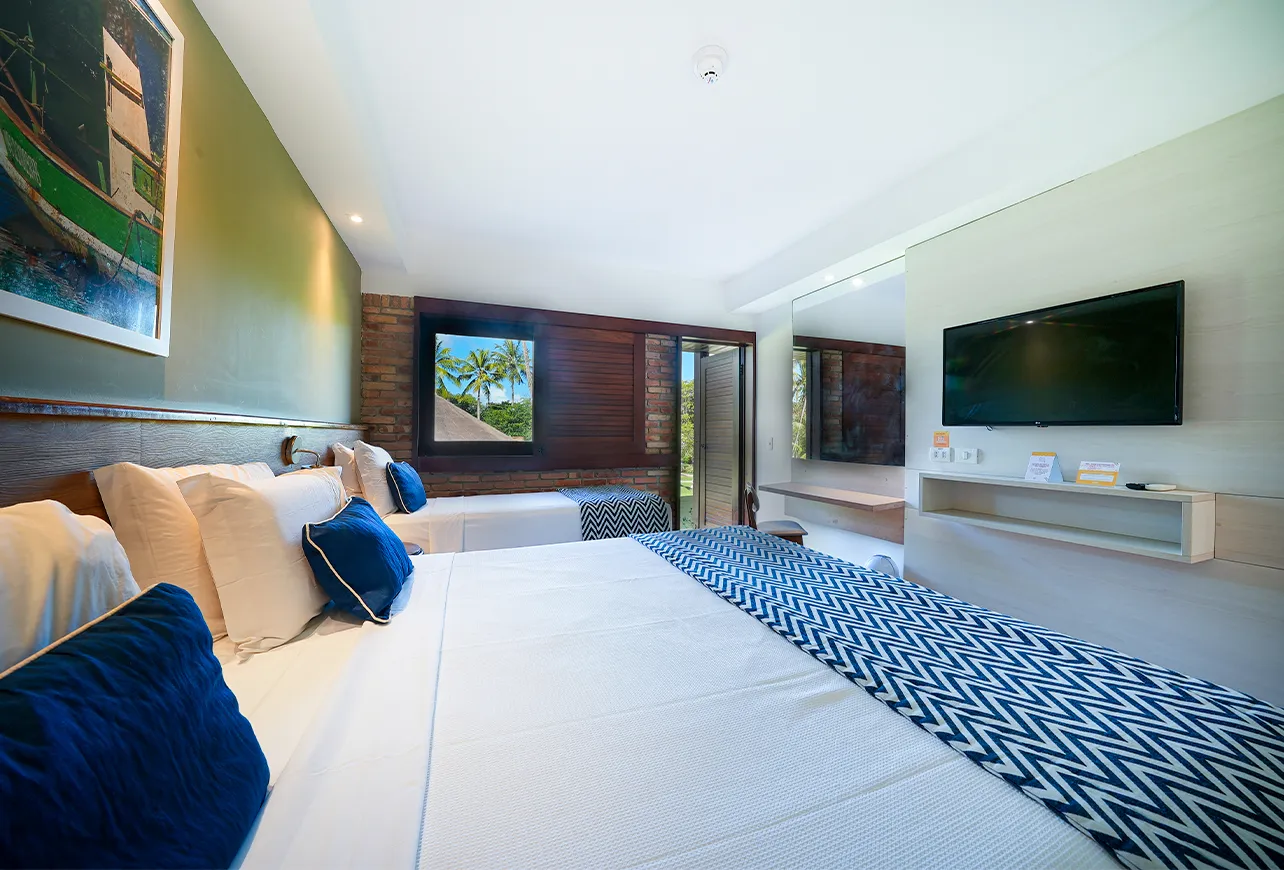 Apartamento Standard, uma das acomodações do Salinas Maragogi, com uma cama queen size e uma cama solteiro. Na frente das camas, tem uma televisão pendurada na parede e, abaixo, uma prateleira de apoio. Ao fundo, a porta de madeira pra varanda com vista pro jardim.