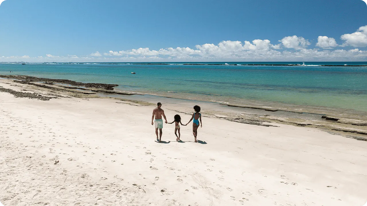 Família caminhando de mãos dadas na areia branca da Praia do Francês com mar e céu azul ao fundo e pequenas algas na faixa de areia.