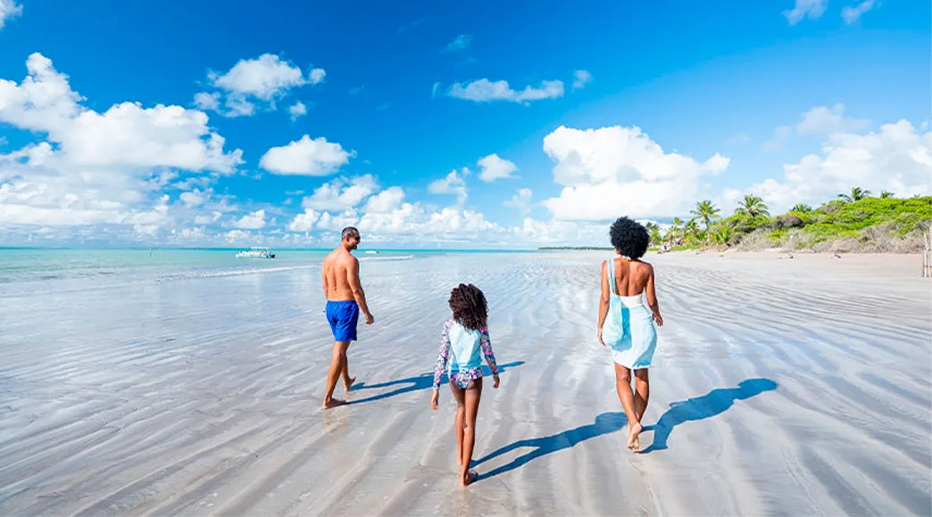 Família com pai, mãe e filha, vistos de costas, caminham pela extensa faixa de areia molhada da Praia de Ipioca durante a maré baixa, sob um céu azul amplo com nuvens.