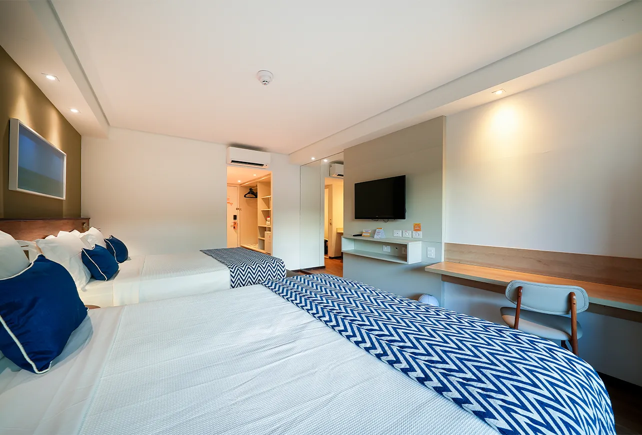 Acomodação Praia Jardim, configuração com duas camas queen size, além de mesa com cadeira, espelhos na parede e roupeiro próximo à porta de entrada. Na frente das camas, uma televisão pendurada na parede com armário de suporte abaixo.
