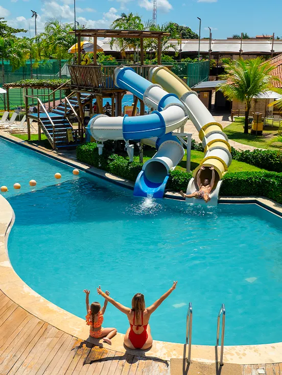 Área de lazer do Salinas Maragogi, com brinquedo de crianças com tobogã colorida na piscina. Família se divertindo, enquanto o pai desce o tobogã, mãe e filha estão sentadas na borda da piscina com os braços pra cima. Ao fundo toda a área com espreguiçadeiras, outros brinquedos, quadras, altos coqueiros verdes e estrutura do resort.