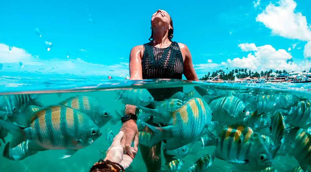 Foto com visão dividida acima e abaixo d'água. Na parte de cima, uma mulher aproveita o sol. Na parte de baixo, um grande cardume de peixes sargentinho nada ao redor de uma mão estendida na água cristalina.