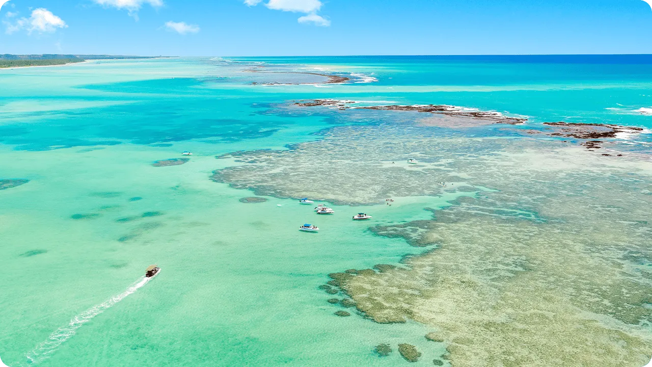 Vista aérea da Praia de Barra Grande com maré baixa e cristalina, recifes de corais visíveis sob a superfície, céu com pequenas nuvens do lado esquerdo e barcos espalhados na água.