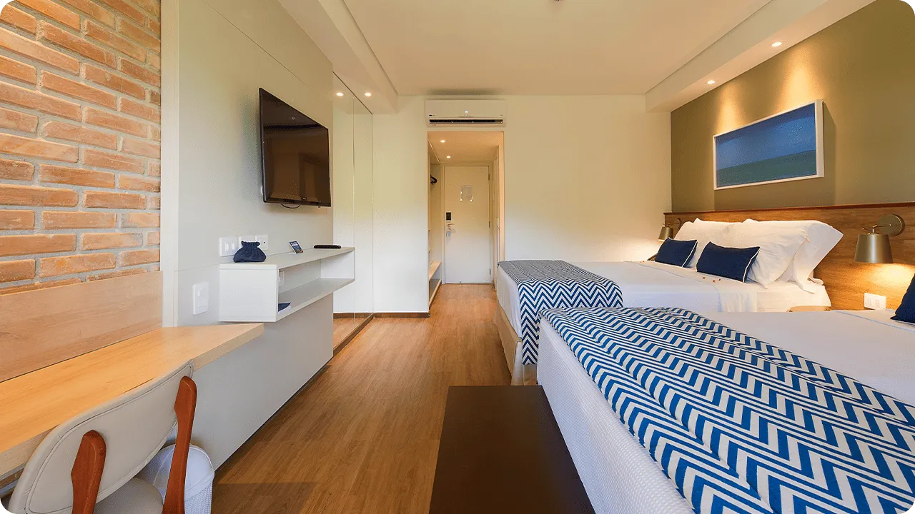 Interior del alojamiento Playa Jardín, con dos camas queen, decorado con imágenes de las playas de Maragogi y paredes de ladrillo, además de una mesa con sillas, espejos, armario y puerta de entrada.
