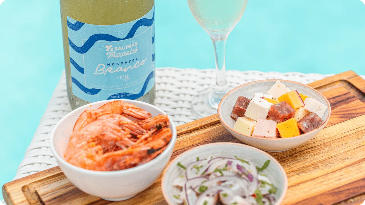 Taça de espumante com vinho moscatel ao lado de petiscos como camarão, frios e cebola temperada, à beira da piscina