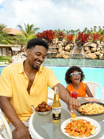 Una familia feliz con padre, madre e hija disfrutando de bocadillos y bebidas en una mesa junto a la piscina en Salinas Maragogi, con camarones, papas fritas y coco.