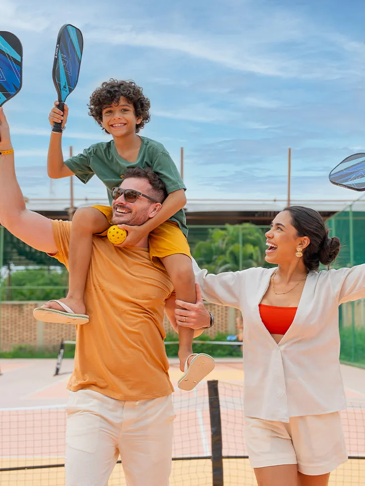 Família na quadra de pickleball, com mãe, pai e filho nas costas do pai. Eles estão com raquetes e bola na mão em área de recreação do Salinas Maragogi.