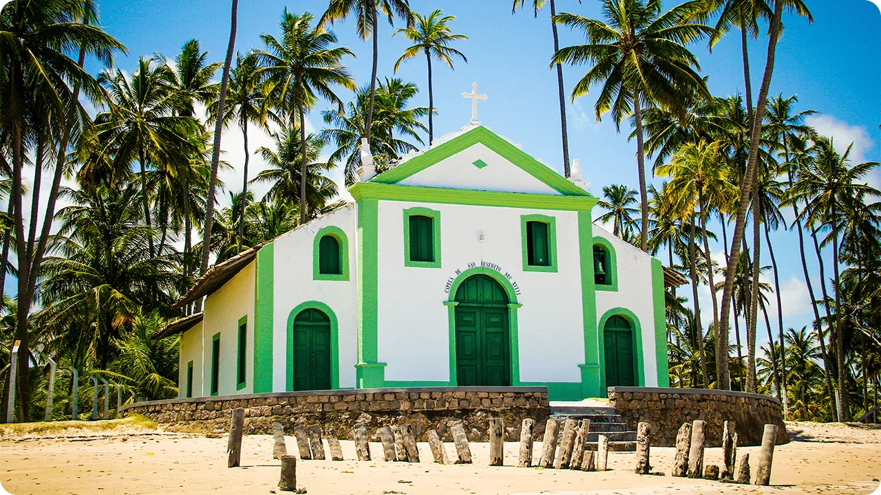 Capela de São Benedito na Praia de Carneiros, com fachada branca e verde, cercada por coqueiros, pequenos tocos de madeira na frente e areia clara sob céu azul