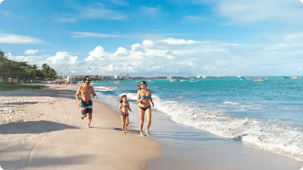 Família correndo pela areia clara da praia de Jatiúca com mar turquesa quase em seus pés, barcos ao fundo e cidade no horizonte.