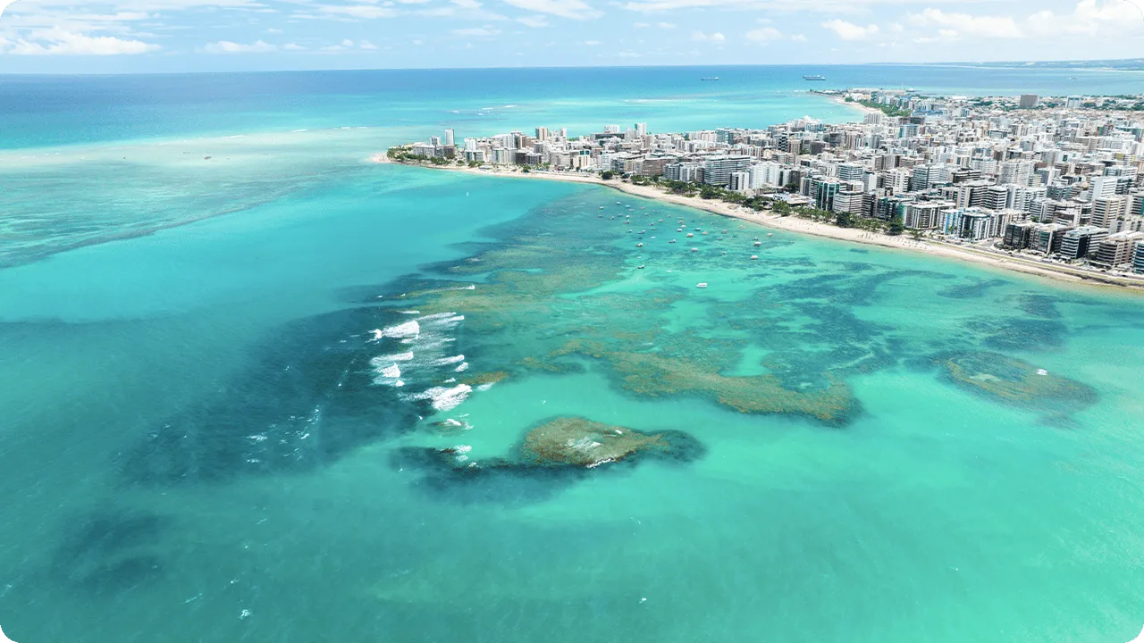 Vista aérea da Praia de Ponta Verde com mar azul cristalino, recifes de corais, pequenos barcos e prédios à beira-mar