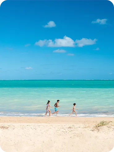 Uma família caminham pela parte rasa do mar turquesa da Praia de Burgalhau, areia clara e céu azul com poucas nuvens a direita.