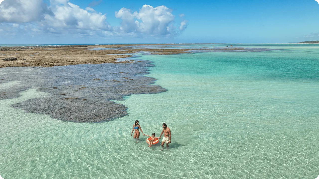 Família com uma criança aproveita as águas rasas e cristalinas da Praia de Ponta de Mangue, cercada por recifes corais aparentes e céu azul com algumas nuvens à esquerda.
