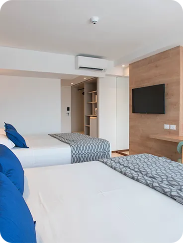 Entrance door to the Deluxe apartment next to the wardrobe, with two queen-size beds placed side by side, two lamps, one for each bed, in front of a wooden panel with cable TV, a desk, and a chair.