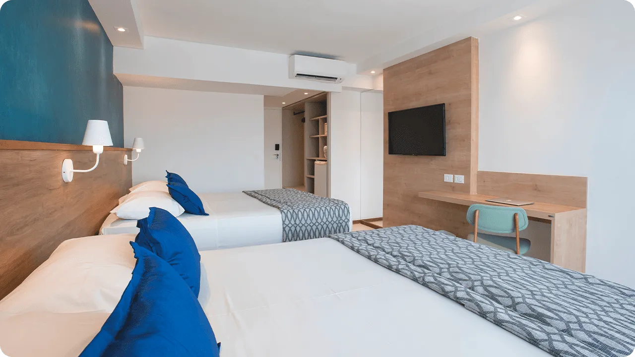 Entrance door to the Deluxe apartment next to the wardrobe, with two queen-size beds placed side by side, two lamps, one for each bed, in front of a wooden panel with cable TV, a desk, and a chair.