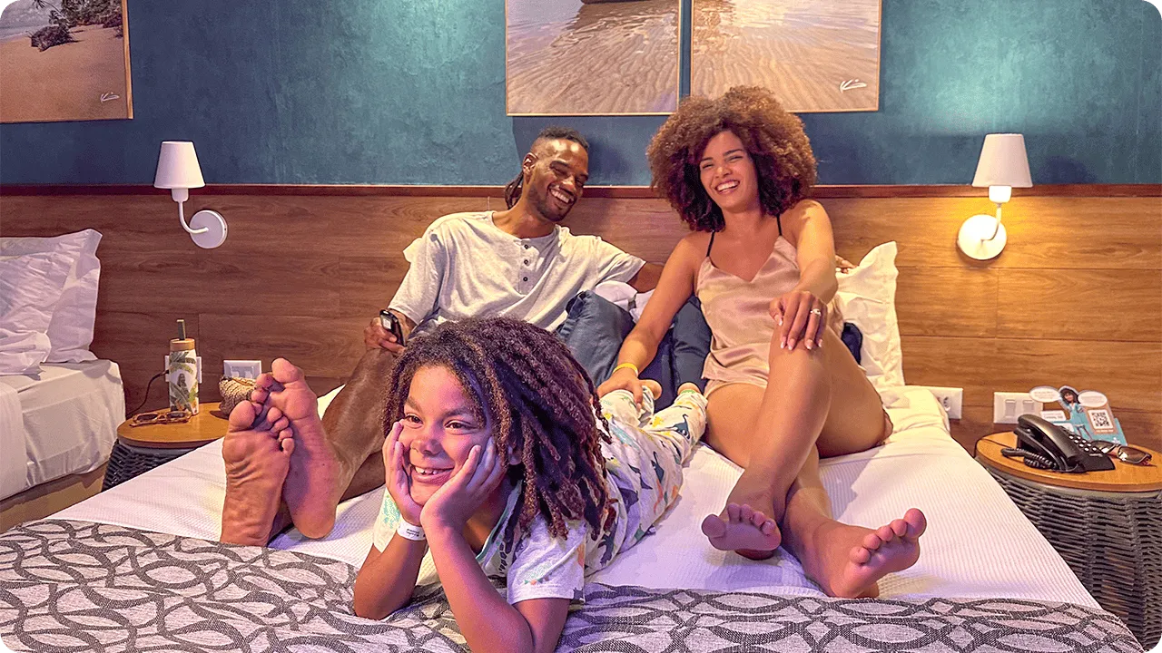 Família feliz relaxando na cama do quarto Standard, com o filho deitado em primeiro plano e os pais sentados atrás. A decoração do quarto inclui uma parede de destaque azul e quadros de praia.