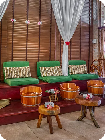 Ambiente de spa com poltronas verdes, baldes de madeira para escalda-pés, decoração com flores tropicais, cortinas brancas e iluminação suave, ideal para momentos de relaxamento e bem-estar.