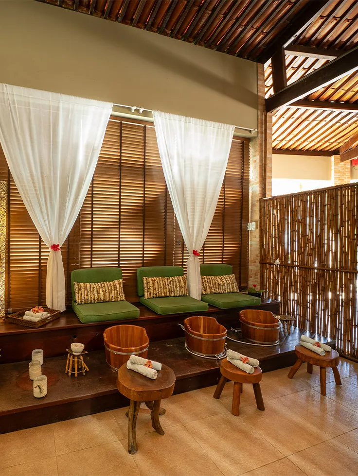 Ambiente de SPA com poofs verdes em cima do banco de madeira, baldes de madeira para escalda-pés, decoração com flores tropicais, cortinas brancas e iluminação suave e uma parede de bambu separando a área do SPA.