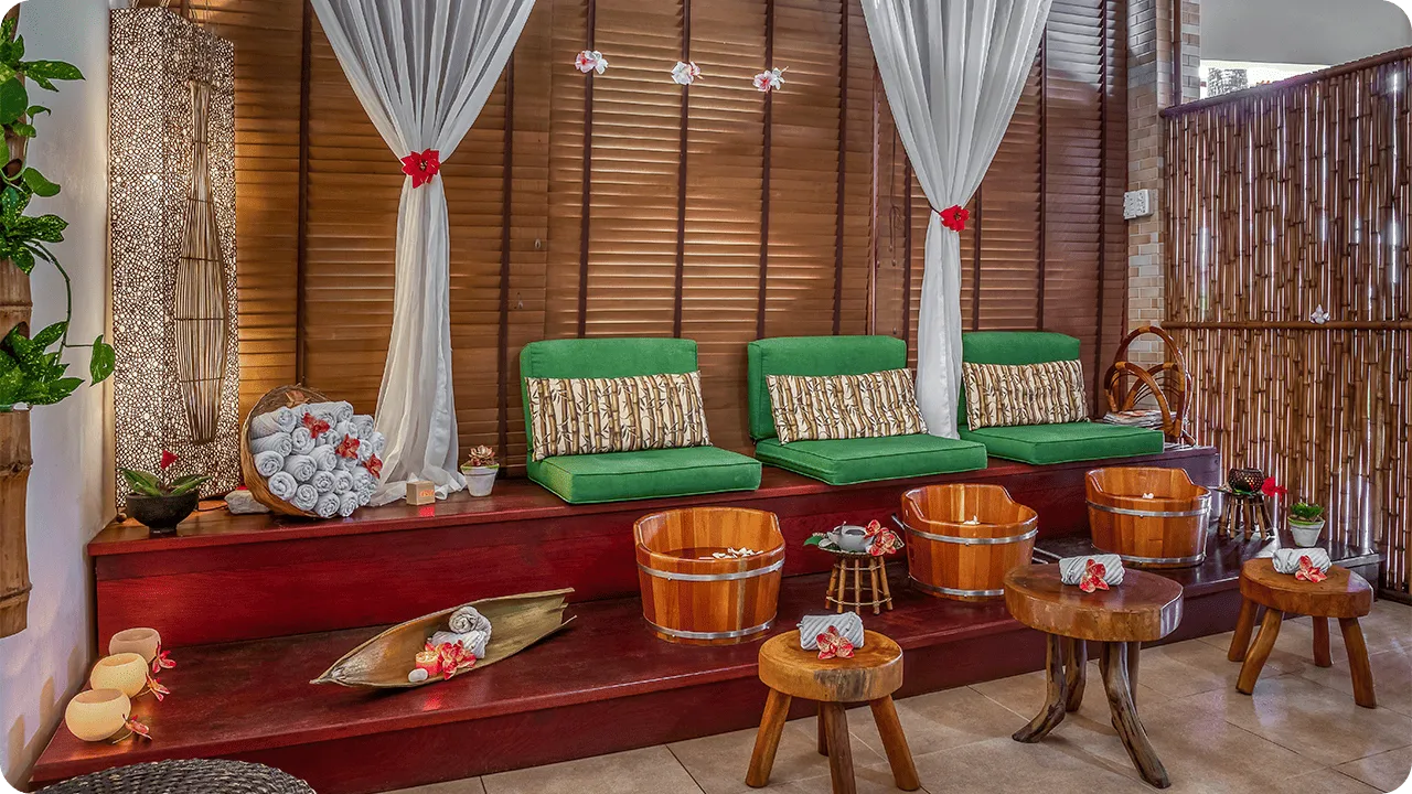 Ambiente de spa com poltronas verdes, baldes de madeira para escalda-pés, decoração com flores tropicais, cortinas brancas e iluminação suave, ideal para momentos de relaxamento e bem-estar.