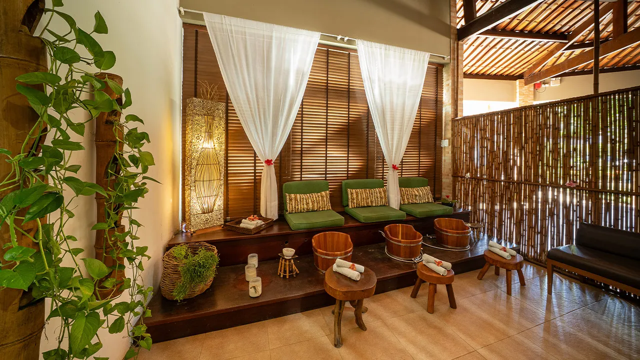 Ambiente de SPA com poofs verdes em cima do banco de madeira, baldes de madeira para escalda-pés, decoração com flores tropicais, cortinas brancas e iluminação suave e uma parede de bambu separando a área do SPA.