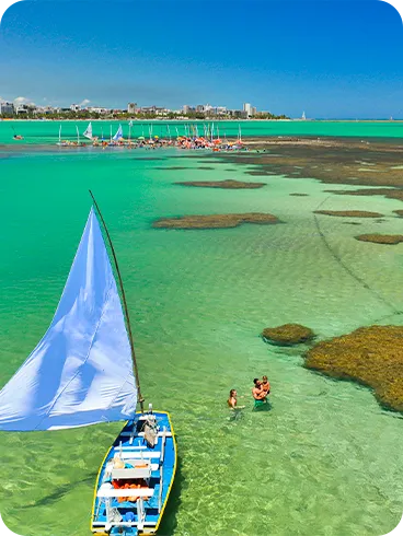 Vista aérea de las piscinas naturales de Pajuçara, Maceió, con una balsa de velas blancas en primer plano navegando por las aguas verde esmeralda. Al fondo, arrecifes de coral y la costa de la ciudad.