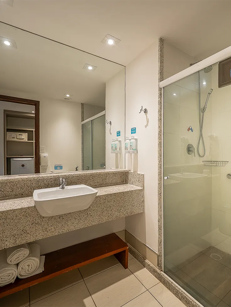 Banheiro do Apartamento Standard no Salinas Maceió com box de vidro, chuveiro, bancada ampla em granito com secador de cabelo pendurado na parede, e um espelho até o teto. 