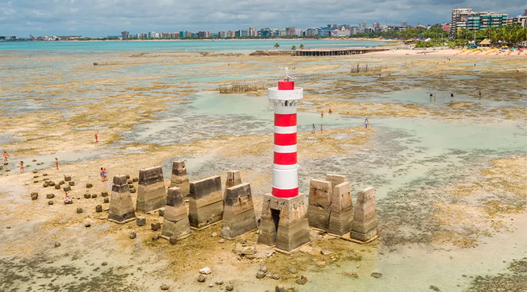 O Farol de Ponta Verde, listrado de vermelho e branco, durante a maré baixa, com o fundo do mar de areia e pedras exposto. Pessoas caminham ao redor da base do farol, com a praia e a cidade de Maceió ao fundo.