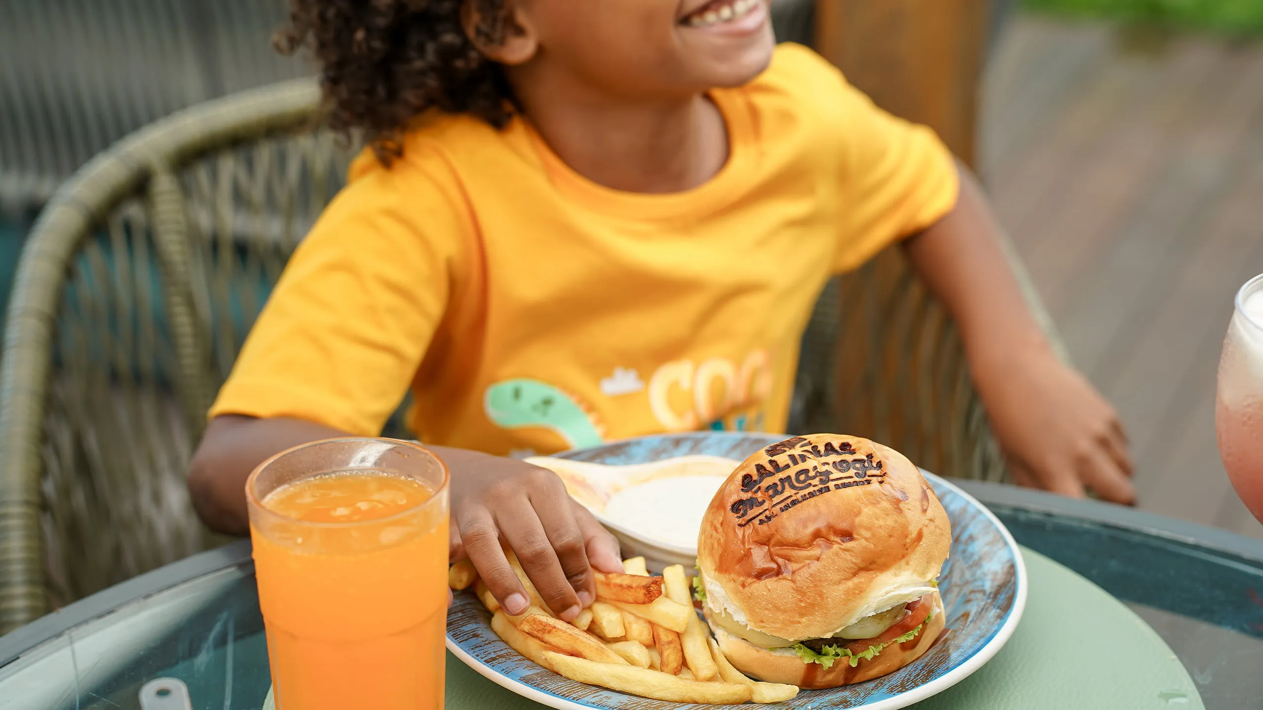 Criança comendo hambúrguer com logo do Salinas Maragogi, batatinha frita e um suco em área externa do resort.