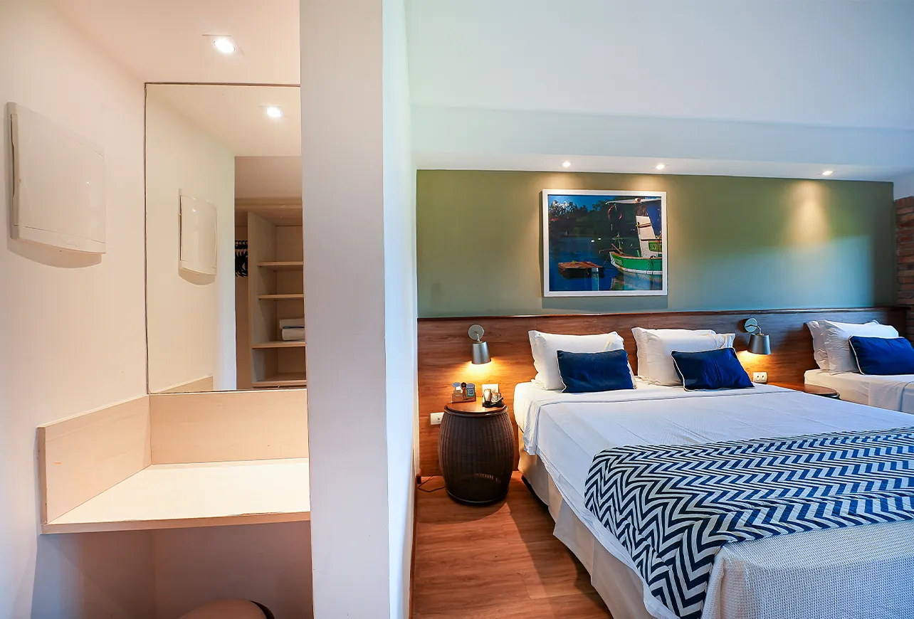 Apartamento Standard, com uma mesa de apoio ao lado esquerdo, como se fosse uma penteadeira com espelho na frente, e ao lado uma cama queen size e uma cama solteiro, chão de madeira e parede verde com quadro pendurado.