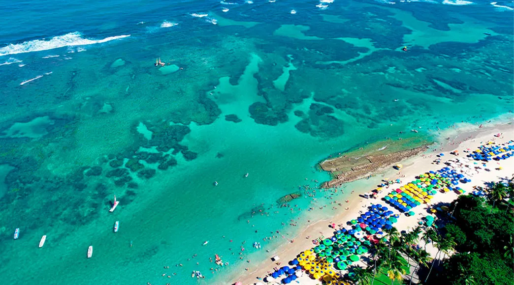 Vista aérea da praia de Porto de Galinhas, mostrando o mar azul-turquesa com seus recifes e a extensa faixa de areia completamente tomada por centenas de guarda-sóis coloridos e banhistas.