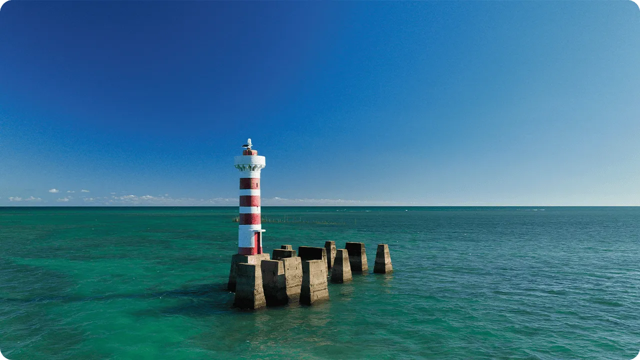 Farol listrado vermelho e branco sobre plataforma de concreto cercado por mar turquesa sob céu azul.