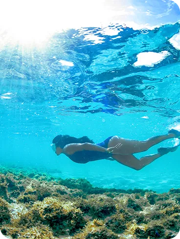 Mulher praticando snorkel em águas tropicais cristalinas, com raios de sol atravessando o mar e fundo coberto por corais.