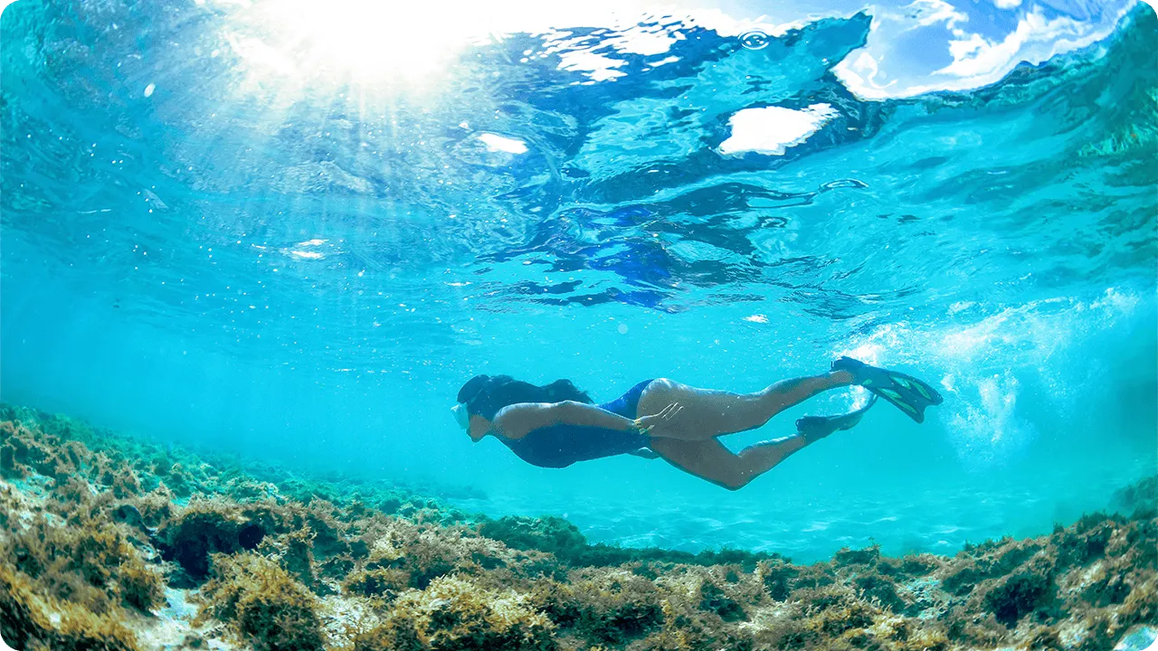 Mulher praticando snorkel em águas tropicais cristalinas, com raios de sol atravessando o mar e fundo coberto por corais.