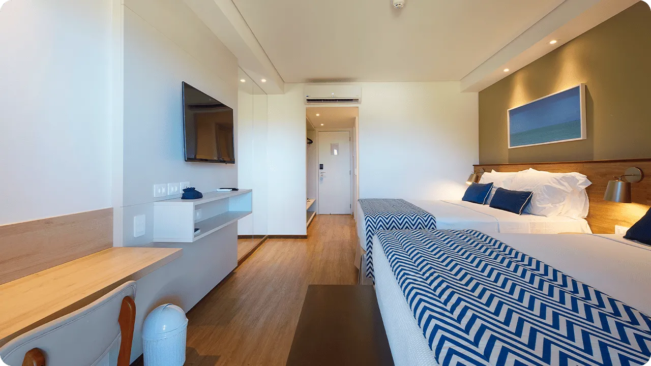 Outro ponto de vista da acomodação, com duas camas queen size, mesa com cadeira, painel com televisão a cabo, decoração aconchegante, quadros com fotografias de praias de Maragogi e porta de entrada.