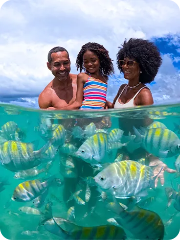 Família sorrindo em mergulho em dia ensolarado na Praia de Pajuçara com peixinhos coloridos nadando em águas turquesa