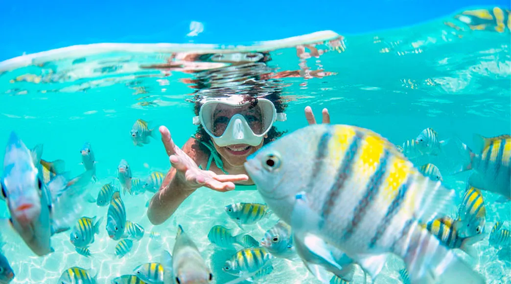 Mulher com máscara de snorkel sorri debaixo d'água, cercada por um grande cardume de peixes sargentinho listrados, em uma piscina natural de água cristalina com fundo de areia visível.