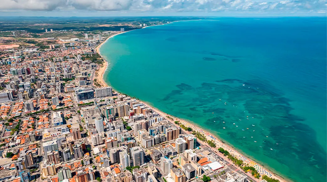Vista aérea panorâmica de toda a orla de Maceió, mostrando a densa cidade com seus prédios de um lado e o mar azul-esverdeado do outro. A longa faixa de praia, incluindo Jatiúca, curva-se ao longo da costa.