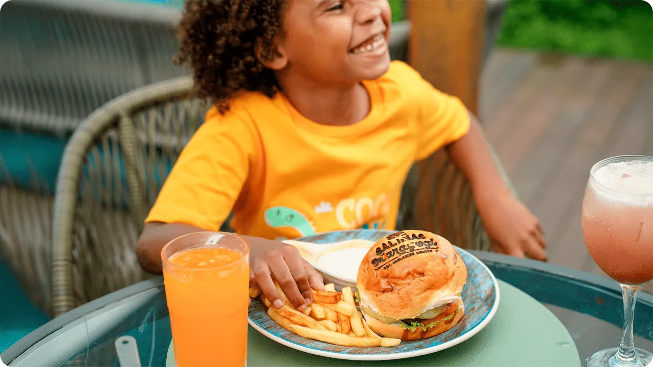 Criança sorridente aproveitando hambúrguer, batata frita e suco durante as férias all inclusive